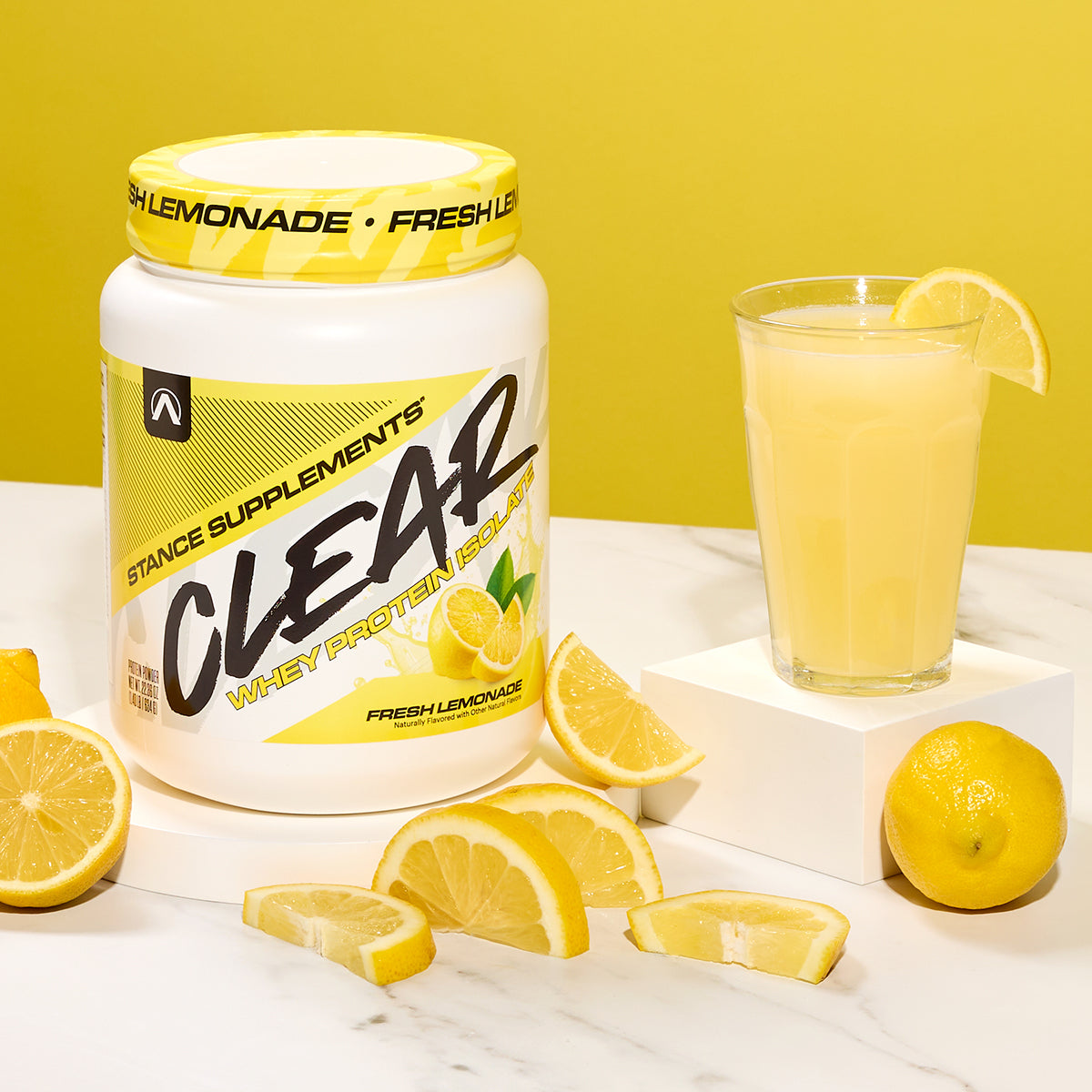 Stance Supplements Clear Whey Protein Powder with a glass of lemonade and lemons on a white surface.